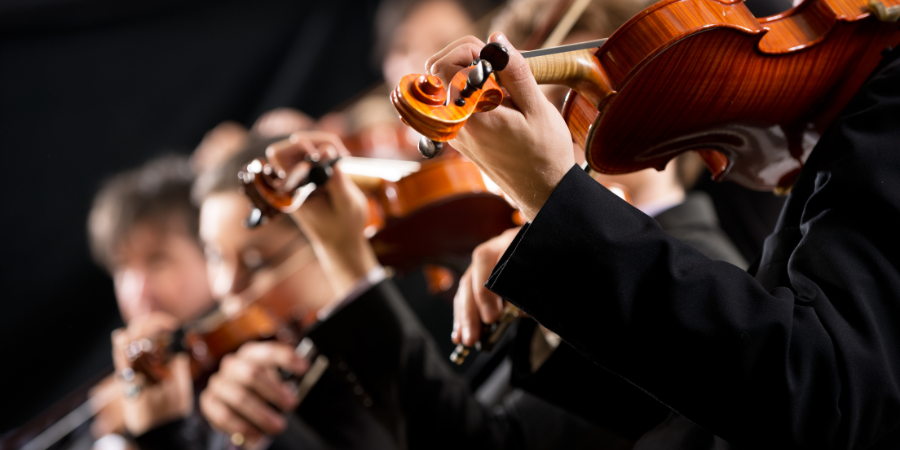 Nahaufnahme mehrerer Geigerinnen und Geiger während einer Aufführung. Im Fokus stehen die Hände und Violinen der Musizierenden, während der Hintergrund unscharf bleibt und die Konzentration sowie das gemeinsame Musizieren im Orchester betont.