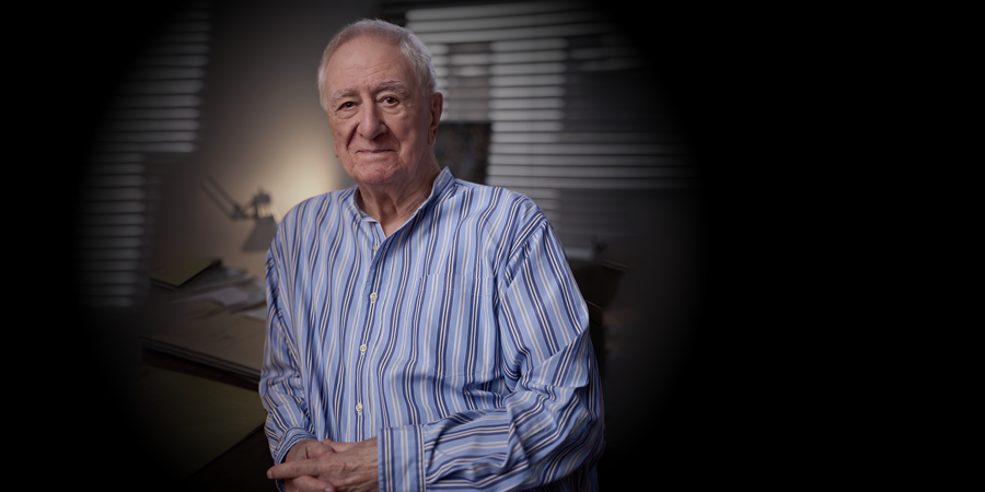 Halbnahaufnahme des Komponisten Bernard Rands in einem blau-weiß gestreiften Hemd. Er sitzt mit gefalteten Händen in einem schwach beleuchteten Büro mit Papieren und Jalousien, umrahmt von einer sanften Vignette.