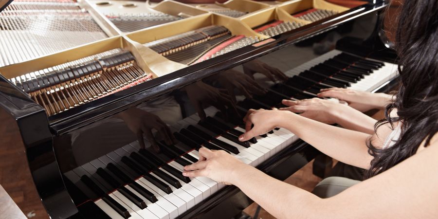 Zwei Personen spielen gemeinsam an einem Flügel vierhändig Klavier; ihre Hände liegen auf den schwarz-weißen Tasten, während im Hintergrund die geöffneten Saiten und Mechaniken des Instruments sichtbar sind.