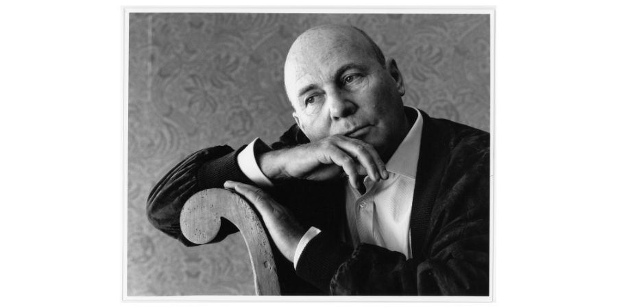 Schwarzweißporträt eines älteren, kahlköpfigen Mannes im Anzug. Es ist Hans Werner Henze. Er lehnt nachdenklich den Kopf auf seine Hand, die auf einer geschwungenen Stuhllehne ruht. Der Blick geht ruhig zur Seite, der Hintergrund ist dezent gemustert.