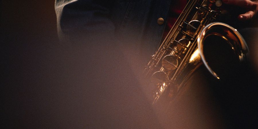 Close-up of a jazz saxophonist playing a golden saxophone, partially obscured by soft focus in the foreground. The musician wears a denim jacket.