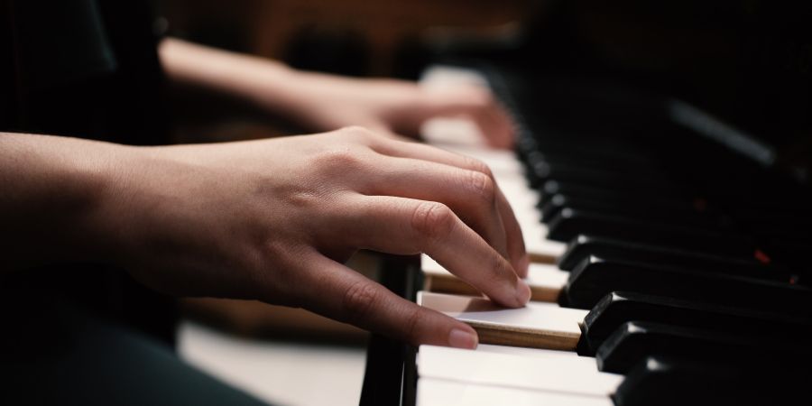Zwei Hände spielen auf einem Klavier. Die Finger liegen auf den Tasten, während der Hintergrund und Teile des Instruments unscharf dargestellt sind. Das Bild konzentriert sich auf die Bewegung und Position der Hände beim Musizieren.
