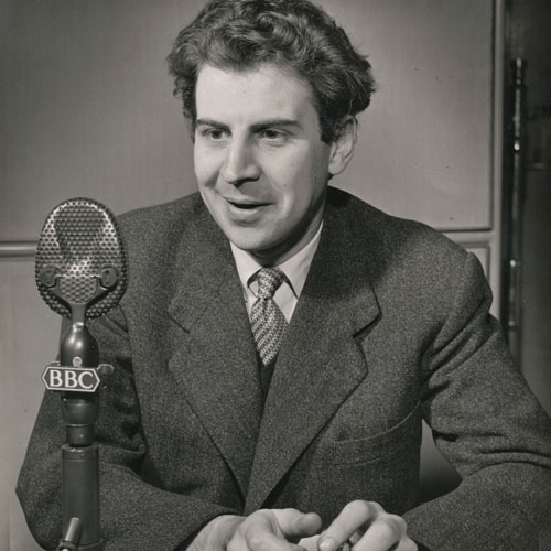 Theodorkias im Studio der BBC London, 1958. Er lächelt freundlich, vor ihm ein Mikrofon, die Hände lose auf dem Tisch verschränkt.