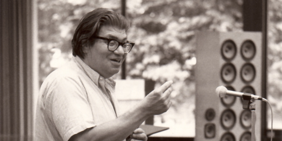 Ein Mann mit Brille und zurückgekämmtem Haar spricht gestikulierend an einem Mikrofon; er steht in einem Innenraum mit Lautsprechern im Hintergrund, die Aufnahme ist in Schwarz-Weiß gehalten.