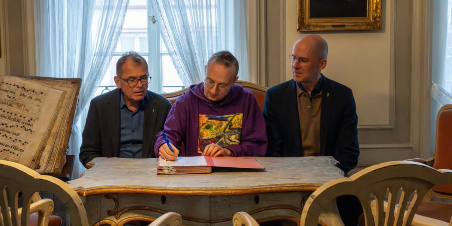 Drei Männer (von links nach rechts: Dr. Thomas Sertl, in der Mitte Alex Paxton, rechts Denis Martin Freiberg) sitzen gemeinsam an einem Holztisch in einem eingerichteten Raum. Der Mann in der Mitte unterschreibt ein Dokument.