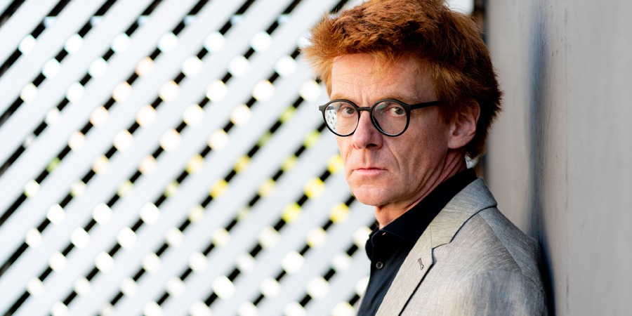 Portrait of Enno Poppe with red hair, round glasses, and a gray blazer, leaning seriously against a wall and looking into the camera; diagonal slats in the background.