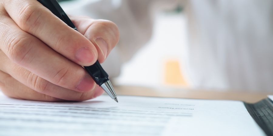 Nahaufnahme einer Hand, die mit einem schwarzen Kugelschreiber ein Dokument unterschreibt. Der Text auf dem Papier ist nur teilweise lesbar.