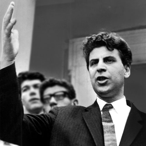 Theodorakis bei einer Rede während der Lambrakis-Friedensbewegung 1963. Er hat die Hand mahnend erhoben, hinter ihm zwei Männer unscharf im Hintergrund.