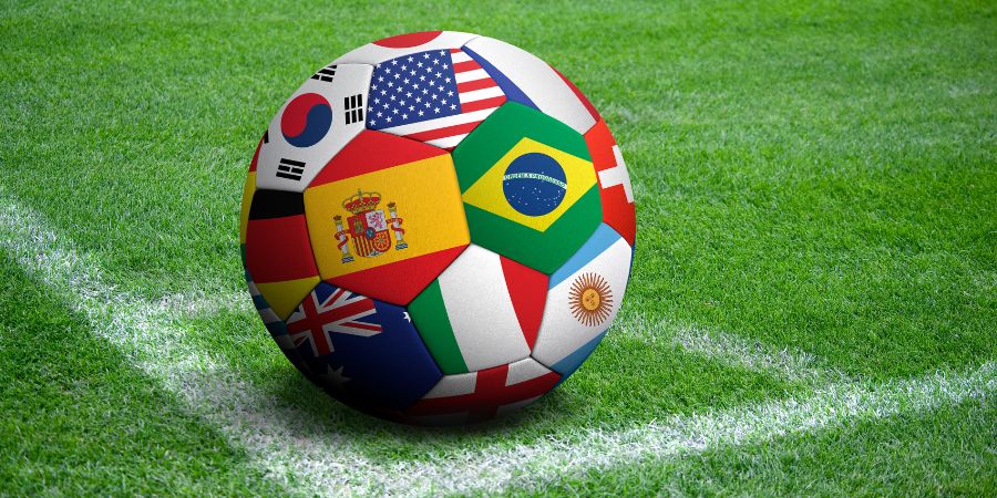 Ein farbenfroher Fußball, dessen Segmente mit Nationalflaggen verschiedener Länder bedruckt sind (darunter Spanien, Brasilien, USA, Südkorea, Argentinien, Deutschland und Italien), liegt auf einem grünen Rasen an der Ecklinie eines Fußballfeldes.