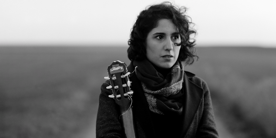Black-and-white portrait of composer and musician Golfam Khayam standing in an open landscape. She gazes into the distance thoughtfully, holding a guitar, with a scarf wrapped around her neck.