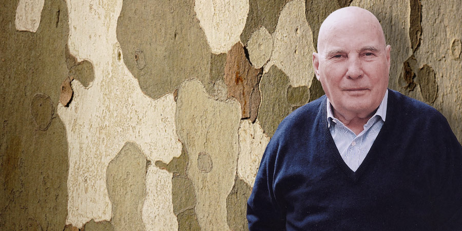Hans Werner Henze in fortgeschrittenem Alter in der rechten Bildhälfte mit offenem Hemdkragen unter dunkelblauem Pullover in die Kamera blickend. Im Hintergrund die Baumrinde einer Platane.