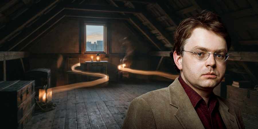 Anno Schreier is on the right side of the frame, wearing a brown jacket and a dark red shirt. He is wearing glasses and looking at the camera. In the background is a dimly lit attic room with a small window offering a view of rooftops beneath a blue eveni