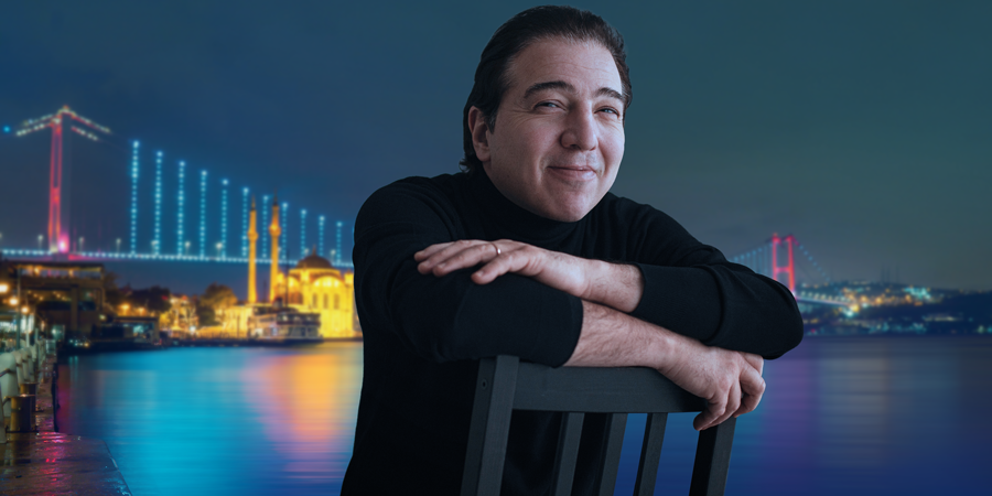 Composer Fazıl Say in the foreground, leaning on a chair against a nocturnal Istanbul backdrop. The illuminated Bosphorus Bridge and Ortaköy Mosque cast shimmering reflections onto the dark waters of the strait.