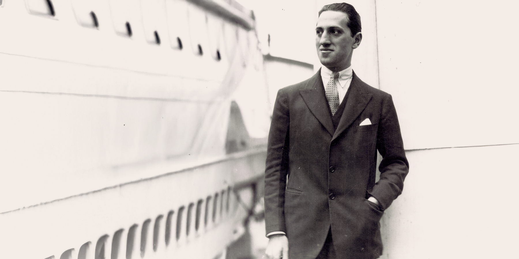 A black-and-white photo of George Gershwin standing by a ship's railing in an elegant suit. Gershwin smiles faintly and gazes to the side, evoking a sense of travel and adventure.