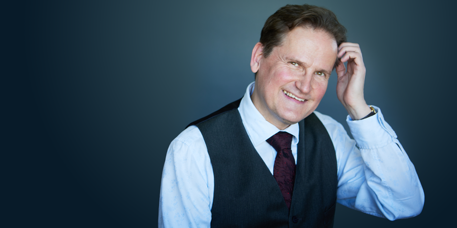 Olli Mustonen, composer and pianist, smiles warmly at the camera. He wears a white shirt, vest, and tie, with one hand touching his head.