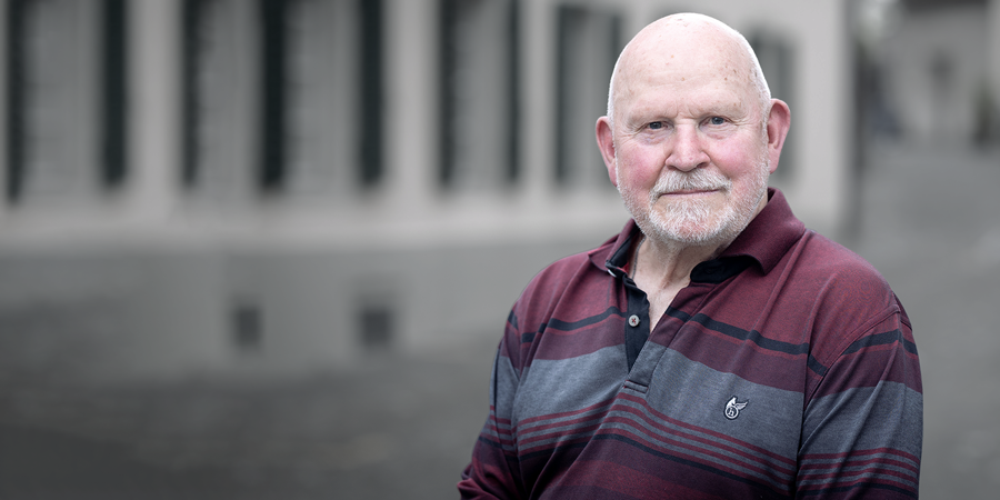 Porträt von Pēteris Vasks: Der Komponist blickt mit einem sanften Lächeln in die Kamera. Er trägt ein weinrot-grau gestreiftes Poloshirt. Der Hintergrund zeigt eine hellgraue Häuserzeile in weicher Unschärfe.