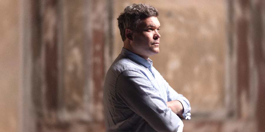 British composer Ryan Wigglesworth stands with arms crossed in front of a blurred, brown-toned wall. He wears a light blue shirt and looks thoughtfully to the side.