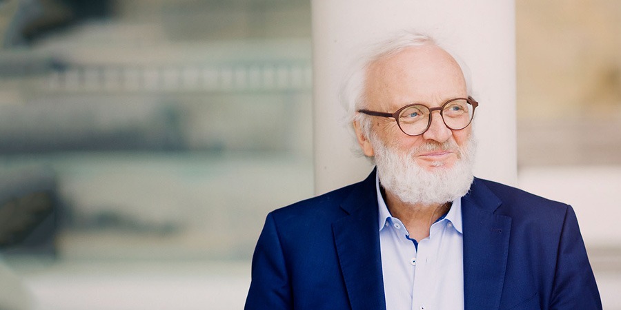 Porträt von Wilfried Hiller: Der Komponist mit weißem Haar und Vollbart blickt lächelnd zur Seite. Er trägt eine Brille, ein hellblaues Hemd und ein dunkelblaues Sakko. Der Hintergrund ist hell und architektonisch-abstrakt gehalten.