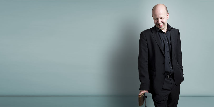Composer Huw Watkins, smiling in a black suit against a gray background. He leans casually on a chair's backrest, with his head tilted slightly to the side.
