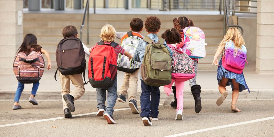 Mehrere Grundschulkinder mit bunten Rucksäcken laufen auf ein Schulgebäude mit Treppen zu.