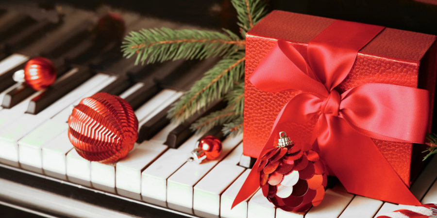 Auf den Tasten eines Klaviers steht eine rote Geschenkbox, die neben einigen dekorativen Kugeln mit unterschiedlichen Texturen auch einen Fichtenzweig enthält, um die weihnachtliche Atmosphäre zu unterstreichen.