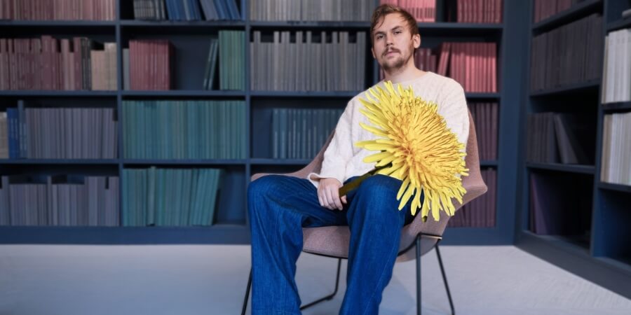 Finnish composer Sebastian Hilli seated in front of color-coordinated bookshelves, holding an oversized yellow flower.