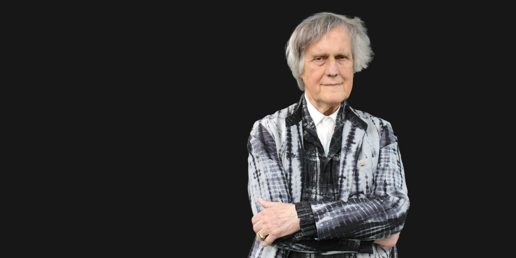 Peter Hanser Strecker vor einem schwarzen Hintergrund. Er trägt ein graues Sakko mit kunstvollen Mustern und Strukturen und lächelt mit verschränkten Armen freundlich in die Kamera