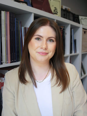 Porträtfoto von Louise Unsworth mit weißem T-Shirt, schmaler goldener Kette und beigem Blazer, freundlich in die Kamera lächelnd. Ein Regal mit Notenbüchern im Hintergrund.