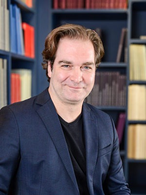 Porträtfoto von Nico Schellhammer mit schwarzem T-Shirt und dunkelblauem Sakko, in die Kamera lächelnd. Im Hintergrund Regale einer Bibliothek.